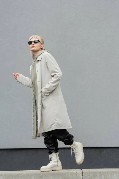 Full length of young woman in fashionable sunglasses, long hoodie, coat, black leather pants and beige boots walking with hand in pocket near grey wall of modern building and looking away — Stock Photo