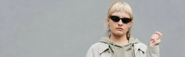 Stylish and young woman with bangs and blonde hair standing in trendy sunglasses and comfortable clothes while looking at camera isolated on grey background in studio, hoodie, banner — Stock Photo