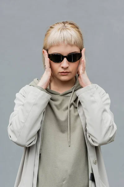 Stylish and young woman with bangs and blonde hair standing in trendy sunglasses and comfortable clothes while looking at camera isolated on grey background in studio, hoodie — Stock Photo