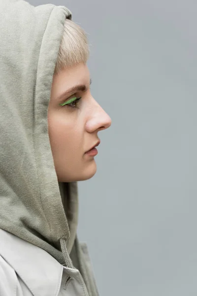 Side view of stylish and young woman with bangs, green eye shadows and blonde hair standing with hood on head and comfortable clothes while looking away isolated on grey background in studio, hoodie — Stock Photo