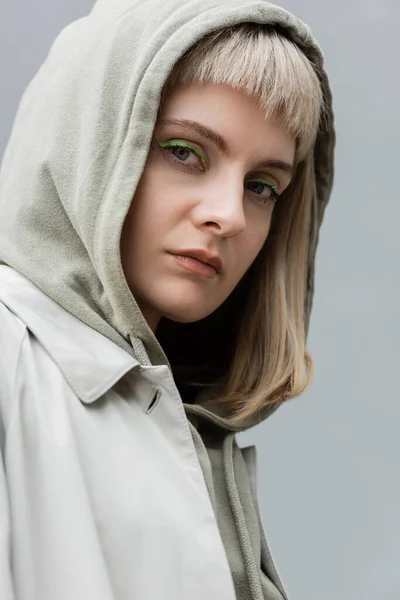 Stylish and young woman with bangs and blonde hair standing with hood on head and trendy comfortable clothes while looking at camera isolated on grey background in studio, hoodie — Stock Photo
