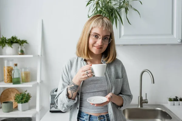 Счастливая молодая женщина с короткими волосами и челкой, очки и татуировки держа чашку утреннего кофе, стоя в повседневной одежде рядом с белыми шкафами и растения в современной кухне — стоковое фото