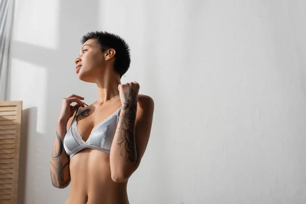 Mujer hipnotizante con pelo corto morena, cuerpo tatuado y pecho sexy con sujetador de seda gris y mirando hacia otro lado mientras está de pie en el dormitorio de luz en casa cerca de la pared blanca - foto de stock