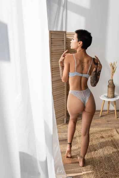 Back view of sultry woman with sexy tattooed body wearing lace panties and bra, standing on wicker rug, looking away near white curtain, room divider and vase with spikelets on bedside table — Stock Photo