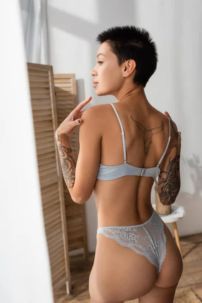 Charming woman with short brunette hair and sexy tattooed body, wearing lace panties and bra, touching shoulders while looking away near room divider in modern bedroom — Stock Photo