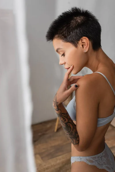 Side view of young captivating woman with short brunette hair and tattooed arm touching face while standing in lace panties and bra near white curtain on blurred foreground in bedroom — Stock Photo