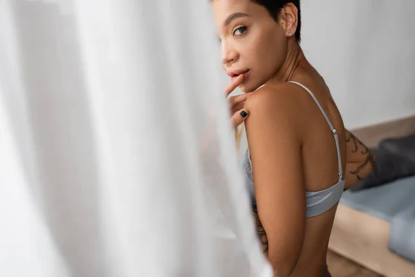 Mujer joven, sexy e intrigante en sujetador tocando los labios y mirando a la cámara mientras está de pie cerca de la cama y la cortina blanca en primer plano borroso en el dormitorio en casa, fotografía boudoir - foto de stock