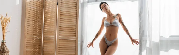 Stunning tattooed woman with short brunette hair and sexy body wearing grey silk lingerie while standing near white curtain, room divider and wicker vase with spikelets in modern bedroom, banner — Stock Photo