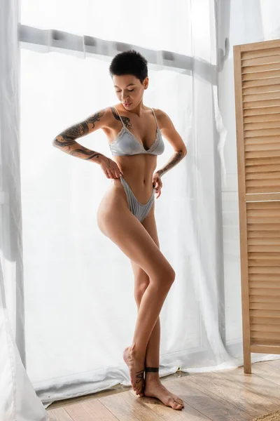 Full length of young and intriguing woman with short brunette hair and sexy tattooed body pulling grey silk panties while standing near white curtain and room divider in modern bedroom — Stock Photo