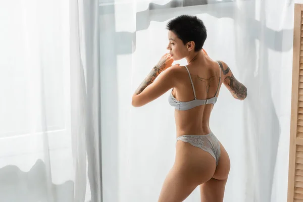Young and stunning woman with sexy tattooed body and short brunette hair wearing lace panties and bra while standing in natural light near white curtain in modern bedroom at home — Stock Photo