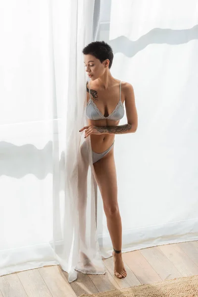 Full length of young, charming and slender woman with sexy tattooed body wearing grey silk lingerie while standing in natural light near white curtain in modern bedroom at home — Stock Photo