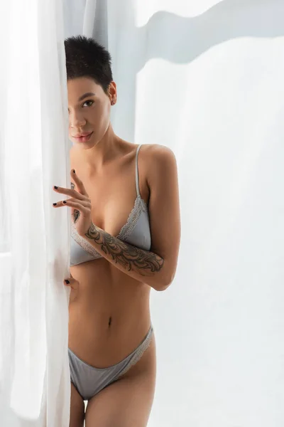 Flirtatious woman with short brunette hair and sexy tattooed body wearing grey silk lingerie such as bra and panties and looking at camera near white curtain in bedroom at home — Stock Photo