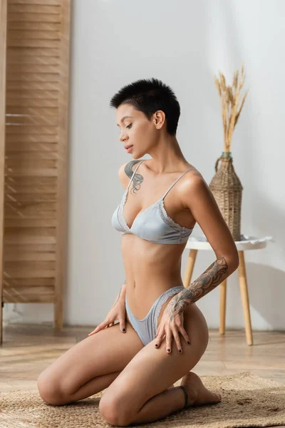 Full length of stunning tattooed woman with sexy body and short brunette hair sitting on wicker rug in lingerie near blurred room divider and vase with spikelets on bedside table in bedroom — Stock Photo
