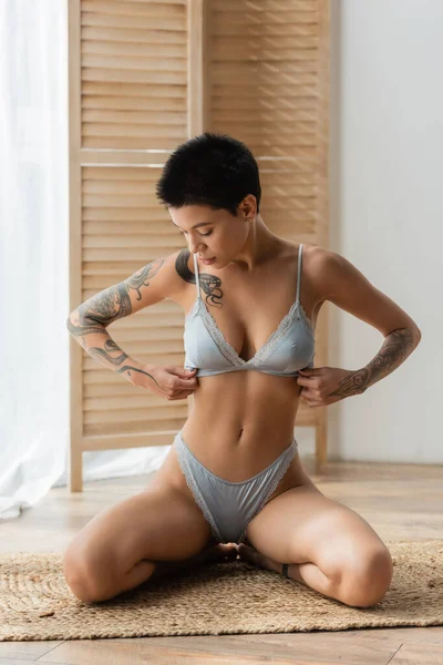 Full length of captivating young woman with short brunette hair and tattooed body adjusting grey silk bra while sitting on wicker rug in seductive pose near room divider on blurred background — Stock Photo