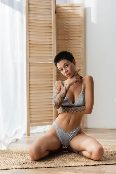 Full length of young passionate woman with short brunette hair and tattooed body wearing grey silk lingerie and sitting in seductive pose on wicker rug near white curtain and room divider in bedroom — Stock Photo