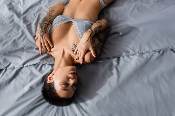 Vista superior de mujer joven y provocativa con pelo corto morena y cuerpo tatuado sexy acostado sobre ropa de cama gris en lencería de seda en el dormitorio moderno en casa - foto de stock