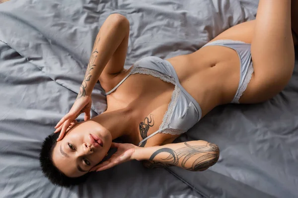 Vista superior da mulher sensual em lingerie, com corpo tatuado sexy e cabelo morena curta tocando o rosto e olhando para a câmera na cama cinza no quarto em casa — Fotografia de Stock