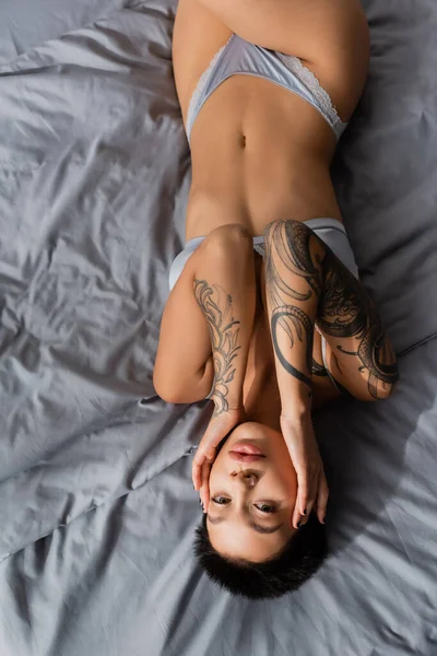 Вид зверху провокаційної молодої жінки з коротким волоссям брюнетки і сексуальним татуйованим тілом, що лежить у спідній білизні на сірій постільній білизні, торкаючись обличчя і дивлячись на камеру — стокове фото