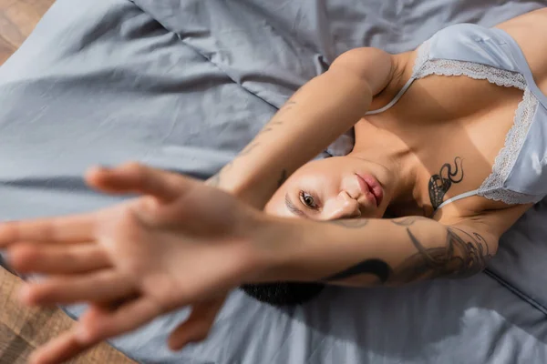 Верхний вид очаровательной и провокационной молодой женщины с сексуальным татуированным телом, лежащим в лифчике с поднятыми руками и смотрящей на камеру на серой кровати на размытом переднем плане — стоковое фото