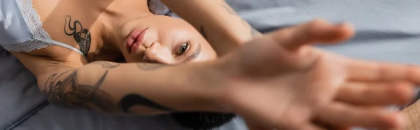 Вид зверху інтригуюча і молода жінка з татуйованим тілом дивиться на камеру, лежачи на сірій постільній білизні з простягнутою рукою на розмитому передньому плані в спальні вдома, банер — стокове фото