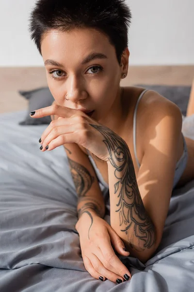Jovem, encantadora e provocante mulher em lingerie, com cabelo curto morena e corpo tatuado sexy segurando a mão perto do rosto e olhando para a câmera em cama cinza em casa — Fotografia de Stock