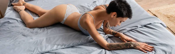 Full length of young, tattooed and passionate woman in lingerie, with sexy body and short brunette hair laying on grey bedding in modern bedroom and home, banner — Stock Photo