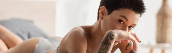 Молодая и соблазнительная женщина с сексуальным татуированным телом и короткими волосами брюнетки, смотрящая в камеру и держась за руку рядом с лицом, лежа в современной спальне дома, будуарная фотография, баннер — стоковое фото