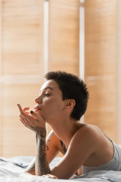 Young, charming and dreamy woman with short brunette hair and sexy tattooed body looking away while laying in bra on white bedding near room divider on blurred background, boudoir photography — Stock Photo