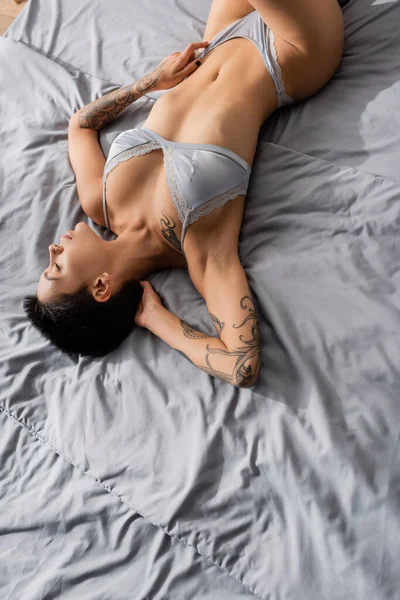 Top view of young and stunning woman in silk lingerie, with sexy tattooed body and short brunette hair laying with closed eyes on grey bedding in modern bedroom, boudoir photography — Stock Photo