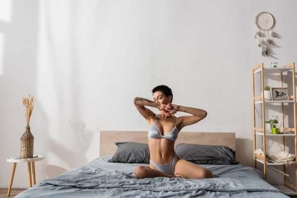 Young seductive woman in silk lingerie, with sexy tattooed body and short brunette hair posing on grey bedding near rack, dream catcher, bedside table and wicker vase with spikelets in bedroom — Stock Photo