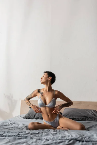 Mujer irresistible en lencería de seda, con pelo corto morena y cuerpo tatuado sexy mirando hacia otro lado mientras posa sobre ropa de cama gris cerca de almohadas en el dormitorio moderno, fotografía boudoir - foto de stock