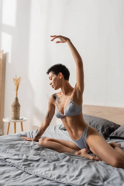 Потрясающая татуированная женщина с сексуальным стройным телом и короткими брюнетками, позирующими с поднятой рукой на серой кровати возле подушек, тумбочкой и вазой с шипами в спальне, будуарной фотографией — стоковое фото