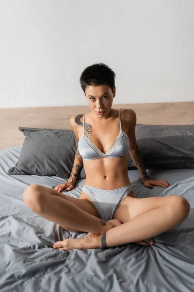 Молодая женщина в шелковом белье, с сексуальным татуированным телом и короткими волосами брюнетки, смотрящей в камеру, сидя в соблазнительной позе со скрещенными ногами на серой кровати возле подушек в спальне — стоковое фото