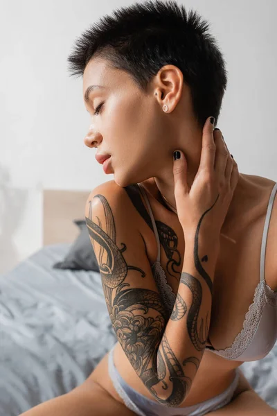 Mujer joven y apasionada en lencería, con cuerpo sexy tatuado, pelo corto morena y ojos cerrados tocando cuello sobre fondo borroso en dormitorio moderno, fotografía boudoir - foto de stock