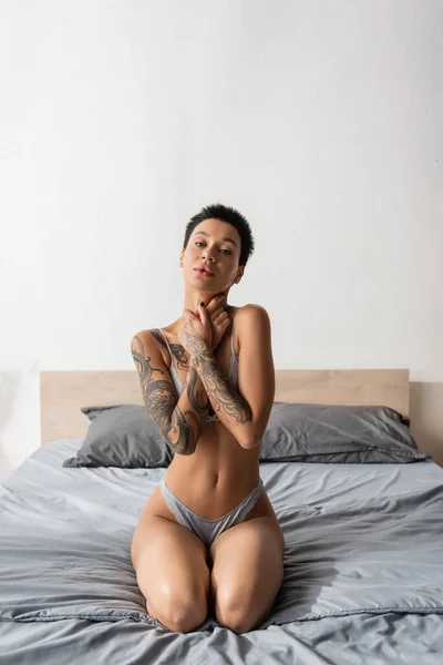 Jovem mulher atraente com cabelo curto morena e corpo tatuado sexy sentado em roupa de cama cinza em lingerie e olhando para a câmera perto de travesseiros no quarto moderno, fotografia boudoir — Fotografia de Stock