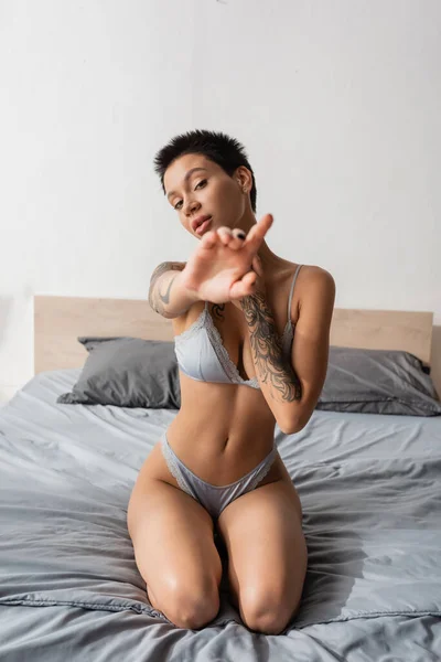 Young and intriguing woman in lingerie, with short brunette hair and sexy tattooed body looking at camera and showing stop gesture on grey bedding near pillows in bedroom, blurred foreground — Stock Photo