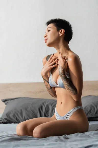 Jeune femme passionnée avec un corps tatoué sexy et des cheveux bruns courts assis sur une literie grise en lingerie de soie et tenant les mains près de la poitrine à côté d'oreillers dans la chambre moderne — Photo de stock