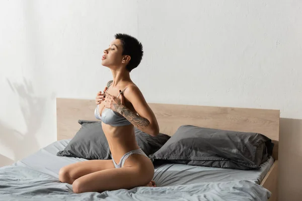Captivating and young woman with short brunette hair and sexy tattooed body touching straps of silk bra while sitting on grey bedding near pillows in modern bedroom at home — Stock Photo