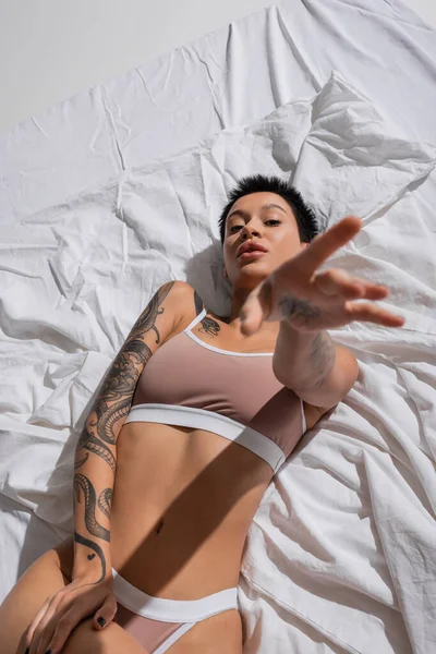 Top view of intriguing young woman in beige lingerie, with short brunette hair and sexy tattooed body laying on white bedding with outstretched hand and looking at camera in studio — Stock Photo