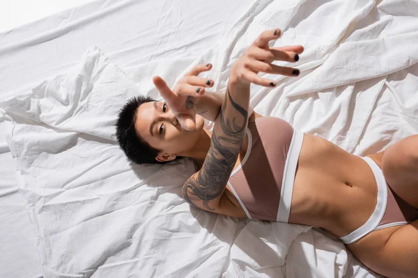 Top view of intriguing tattooed woman in beige lingerie, with sexy body and short brunette hair laying on white bedding, pointing with fingers and looking at camera in studio, erotic photography — Stock Photo