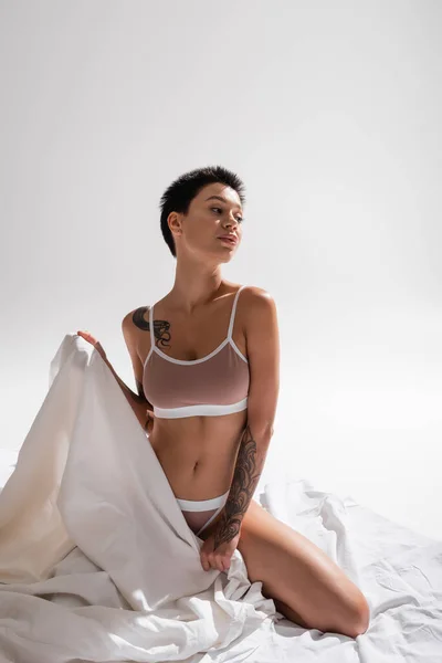 Jeune femme tatouée et passionnée en lingerie beige, avec corps sexy et cheveux bruns courts tenant un drap de lit blanc et regardant loin en studio sur fond gris, photographie érotique — Photo de stock