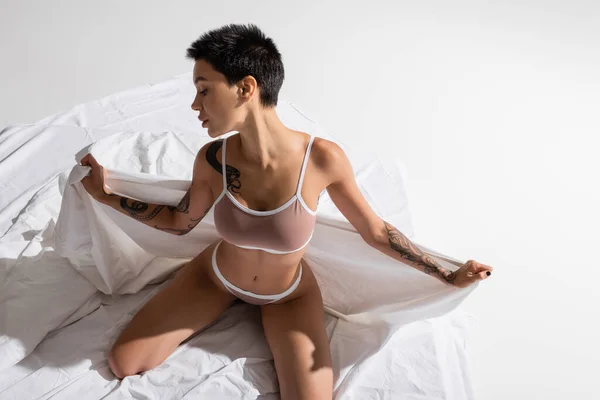 High angle view of young and seductive woman in beige lingerie, with short brunette hair and sexy tattooed body posing on white bedding and grey background, art of seduction — Stock Photo