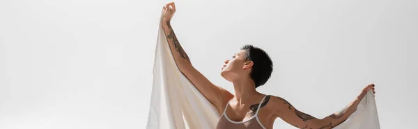 Jeune et séduisante femme en soutien-gorge beige, avec un corps tatoué sexy et des cheveux bruns courts tenant un drap de lit blanc tout en posant sur fond gris en studio, bannière — Photo de stock