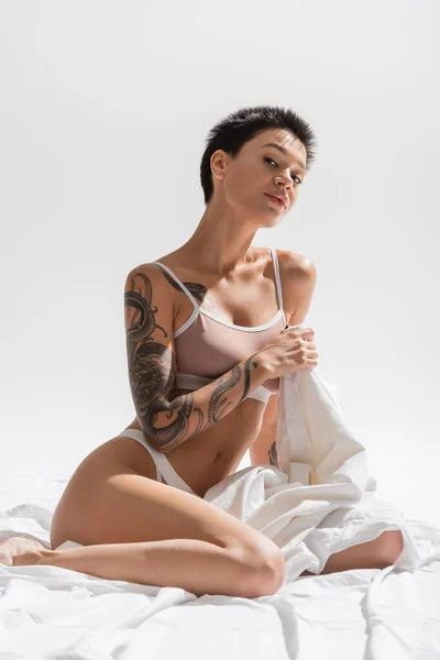 Joven mujer coqueta en sujetador beige y bragas, con cuerpo tatuado sexy y pelo corto morena sentado con sábana blanca y mirando a la cámara sobre fondo gris, fotografía erótica - foto de stock