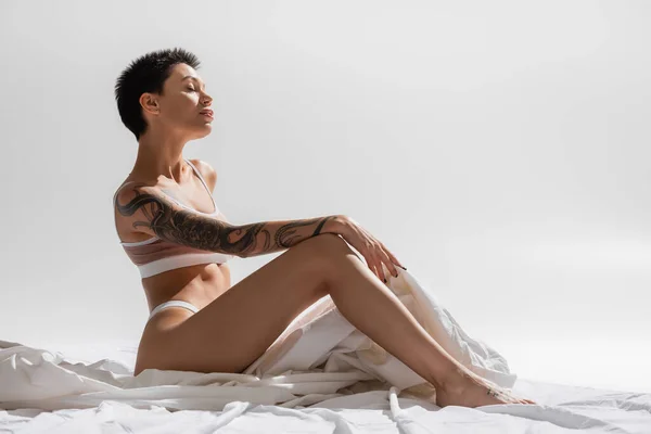 Vista lateral de mujer joven agraciada con cuerpo tatuado sexy y pelo corto morena sentado en lencería beige con los ojos cerrados sobre ropa de cama blanca y fondo gris en el estudio, fotografía erótica - foto de stock