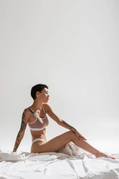 Mujer joven, cautivadora y tatuada con el pelo corto morena sentado en la ropa de cama blanca y tocando la pierna delgada sobre fondo gris con espacio de copia en el estudio, fotografía erótica - foto de stock