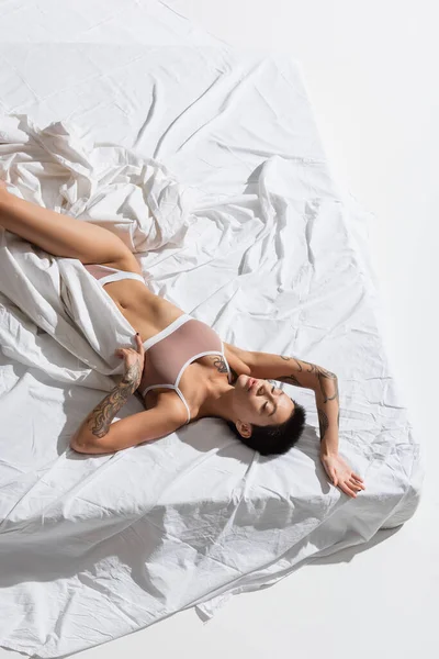 Top view of stunning and slender woman in beige lingerie, with short brunette hair and sexy tattooed body laying with closed eyes on white bedding in studio, erotic photography — Stock Photo
