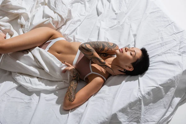 Top view of young and passionate woman with closed eyes, sexy tattooed body and short brunette hair touching neck while laying on grey bedding in studio, erotic photography — Stock Photo