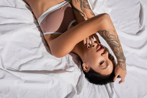 Vista superior da mulher jovem e encantadora com cabelo curto morena e corpo tatuado sexy deitado em lingerie bege em roupa de cama branca e olhando para a câmera, arte da sedução — Fotografia de Stock