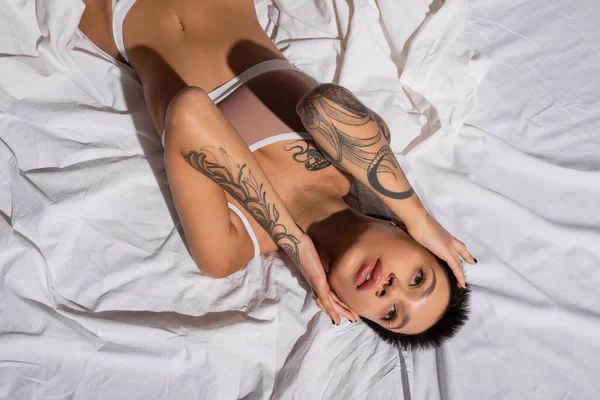 Top view of charming and passionate young woman in beige lingerie, with sexy tattooed body and short brunette hair touching face and looking away while laying on white bedding — Stock Photo